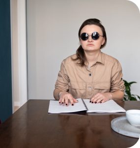 На фото слепая девушка сидит за столом и читает книгу с шрифтом Брайля